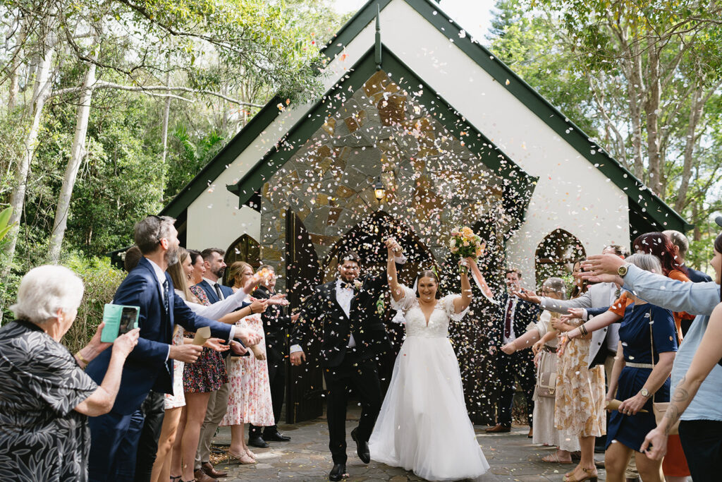 Courtney and Luke walking through confetti thrown by their guests. The images is taken in front of Coolibah Downs Private Estate stone chapel. 