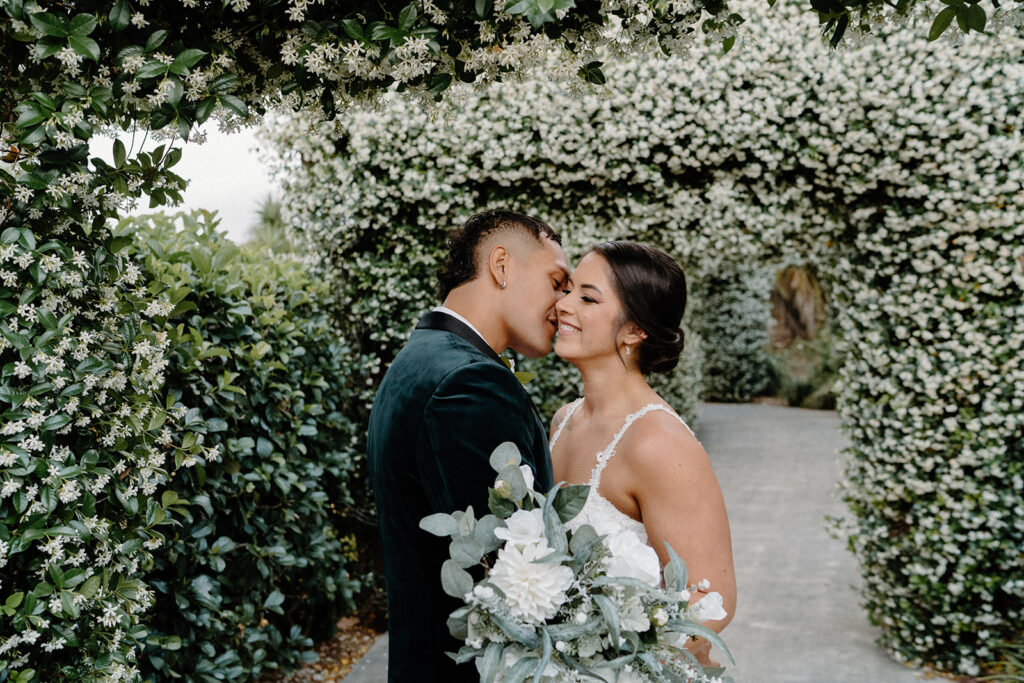 A bride and groom kissing in The Club Parkwood's jasmine archway toward which leads toward their wedding ceremony. 